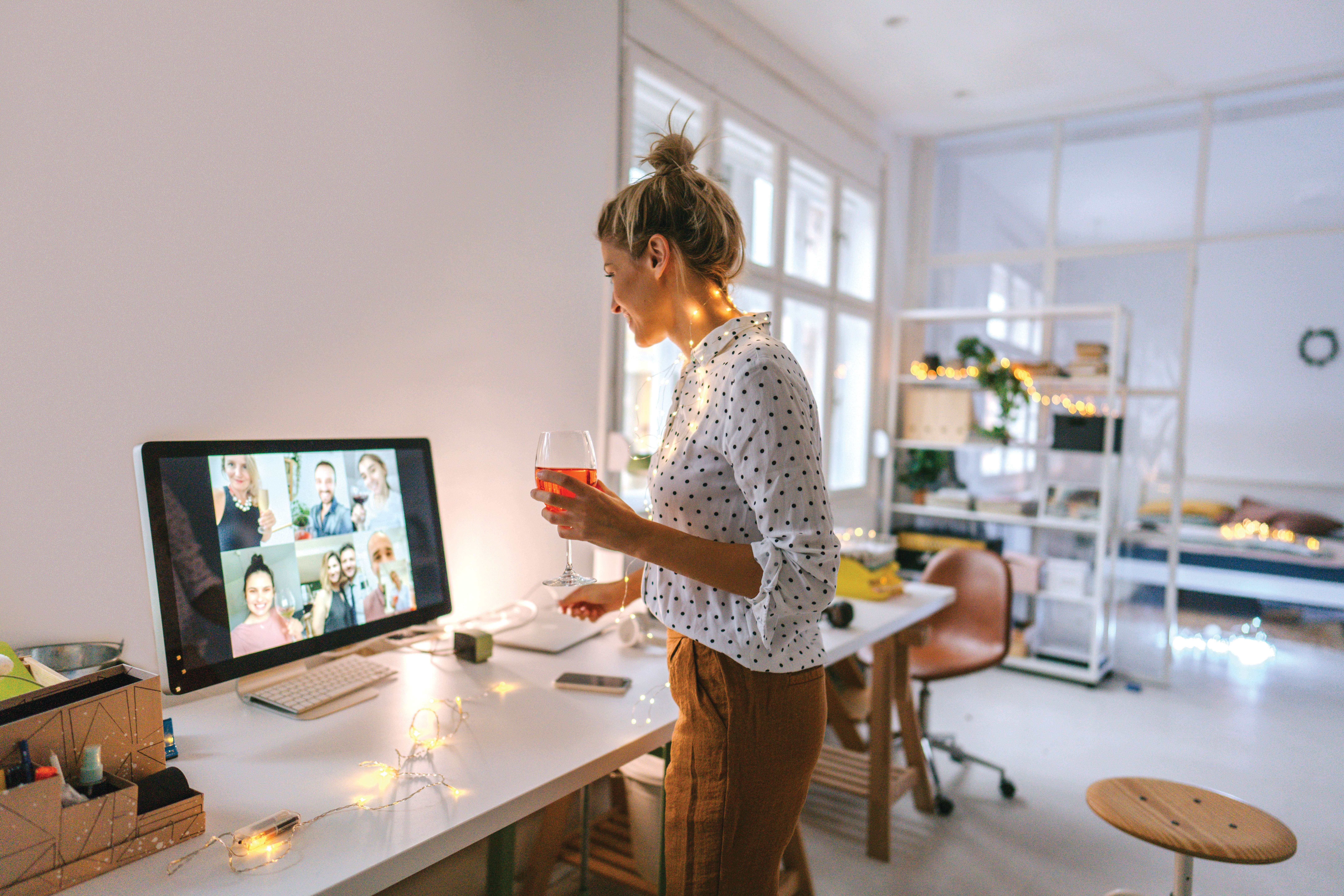 Photo of a young woman having a virtual happy hour with her friends; enjoying a glass of wine in the living room of her apartment while having a video call with a group of friends.