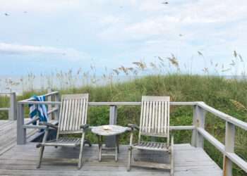 Rustic porch looking out over sand dunes and ocean. Towel hanging on the railing of porch deck.