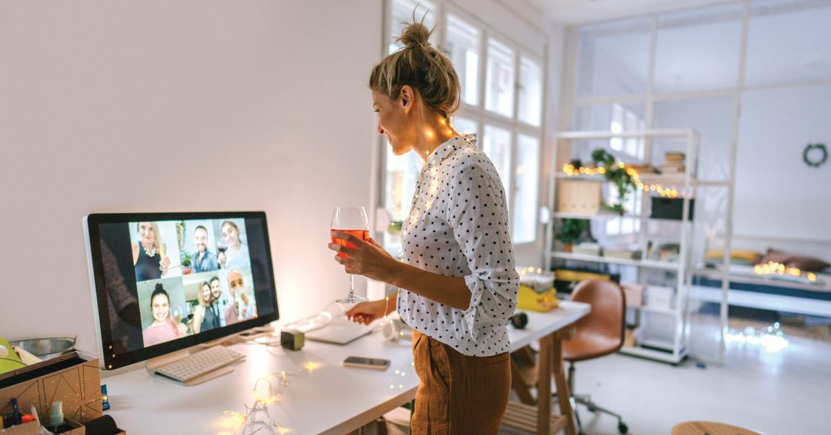 Photo of a young woman having a virtual happy hour with her friends; enjoying a glass of wine in the living room of her apartment while having a video call with a group of friends.