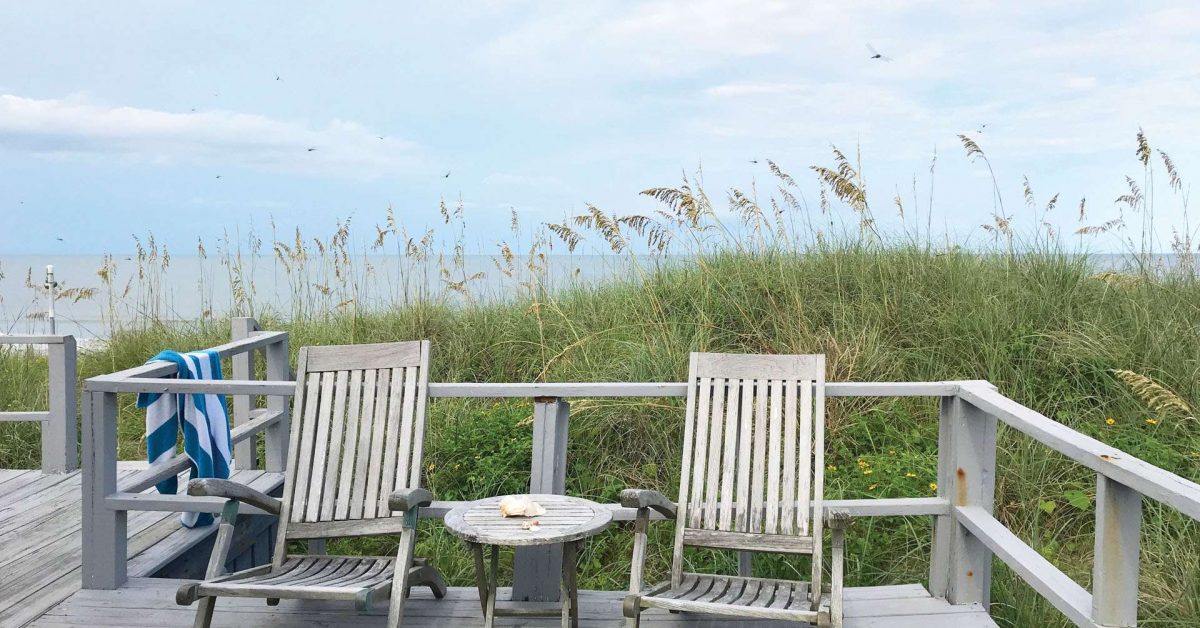 Rustic porch looking out over sand dunes and ocean. Towel hanging on the railing of porch deck.