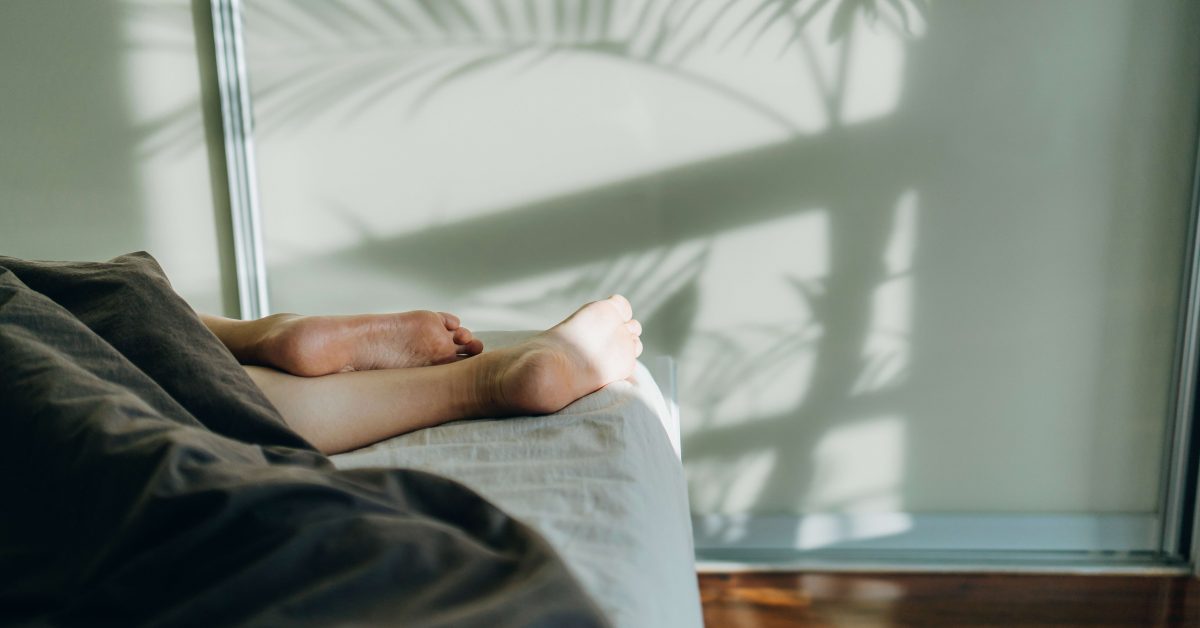 Low section of young woman sleeping in bed, with bare feet under the blanket in bedroom. Sunlight shining through window with the silhouette of palm tree on a beautiful sunny morning